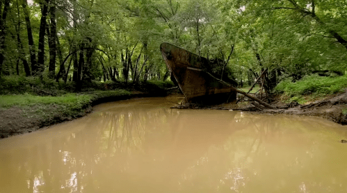 An abandoned shipwreck rests in a muddy creek, surrounded by lush green trees.
