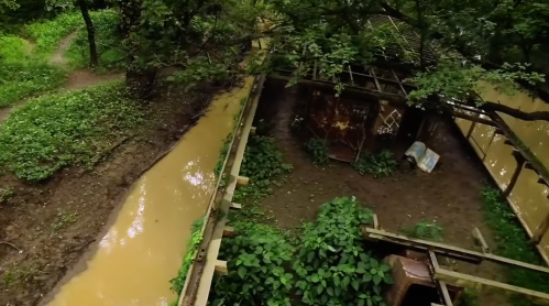 Aerial view of a lush green area with a muddy stream and a dilapidated structure surrounded by trees.