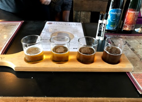 A wooden flight paddle holds four small glasses of different beers, with menus and drinks in the background.