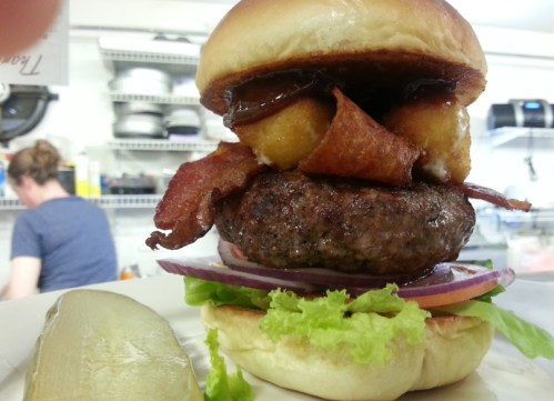 A towering burger with a beef patty, bacon, onion, lettuce, and a crispy topping, served with a pickle on the side.