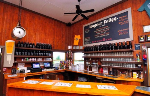 A rustic bar with wooden walls, a long counter, beer taps, and a menu board displaying drinks and tastings.