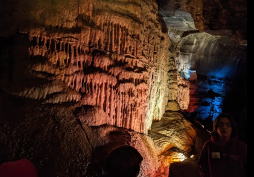 Stalactites and stalagmites illuminated in a cave, with colorful lighting and a person observing nearby.