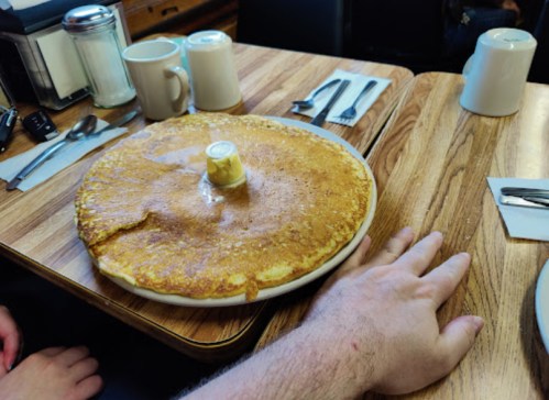 A large pancake with a pat of butter in the center sits on a table next to a hand, coffee cups, and utensils.