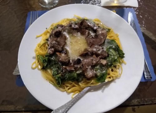 A plate of pasta topped with sautéed mushrooms, greens, and sliced meat, garnished with cheese.