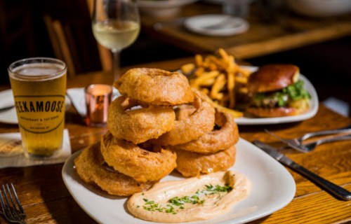 A plate of stacked onion rings with dipping sauce, alongside a burger and fries, with a glass of beer in the background.