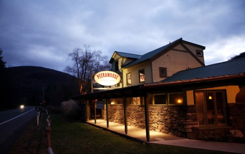 A cozy restaurant named "Peekamoose" illuminated at dusk, surrounded by trees and mountains.