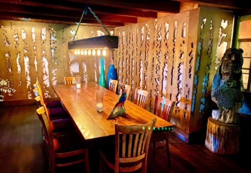 A rustic dining room with a long wooden table, wooden chairs, and artistic wall cutouts, illuminated by pendant lights.