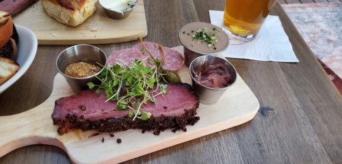 A wooden board with sliced meat, garnished with microgreens, mustard, pickles, and a side of beer on a table.