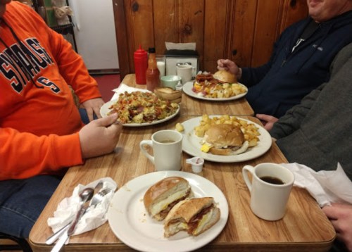 Three people enjoy a hearty meal at a wooden table, featuring plates of food and cups of coffee.