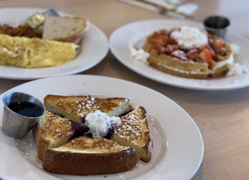 A plate of French toast with syrup, an omelet, and a waffle topped with fruit and whipped cream.