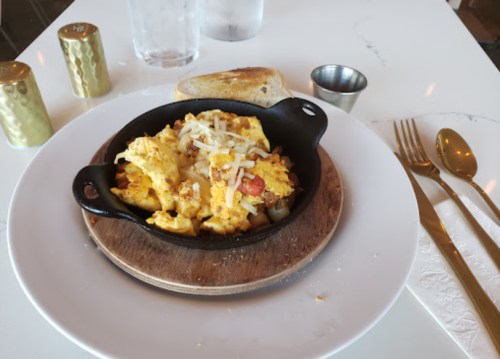 A skillet of cheesy scrambled eggs with diced tomatoes and onions, served with toast and utensils on a white table.