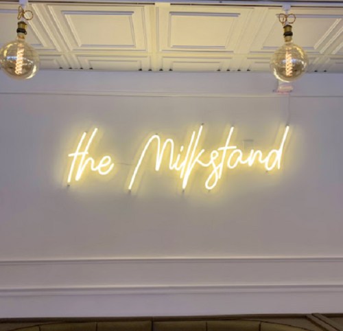 Neon sign reading "the Milkstand" illuminated against a white wall with decorative ceiling.