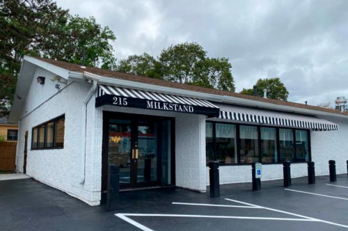 Exterior of a white building with a striped awning labeled "MILKSTAND," featuring large windows and a paved parking area.