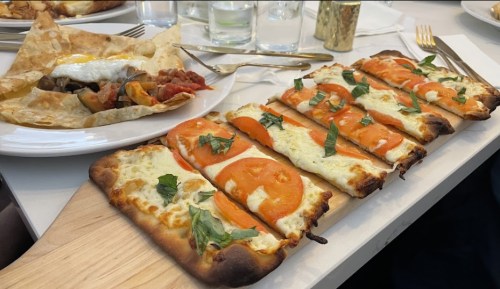 A wooden board with flatbread topped with cheese, tomatoes, and basil, alongside a plate of vegetables and an egg.