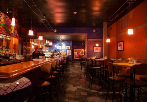 A cozy restaurant interior with a bar, colorful walls, and empty tables under warm lighting.