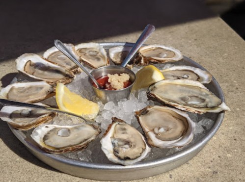 A platter of fresh oysters on ice, served with lemon wedges and a small bowl of cocktail sauce.