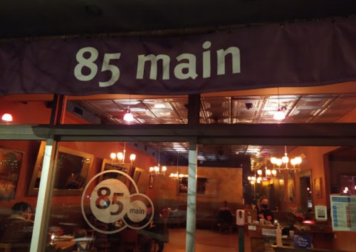 Sign for "85 Main" above a restaurant entrance, with warm lighting and patrons visible inside.