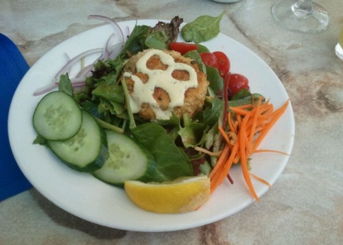 A plate of fresh salad with greens, cherry tomatoes, cucumber, carrots, and a fried patty drizzled with sauce.