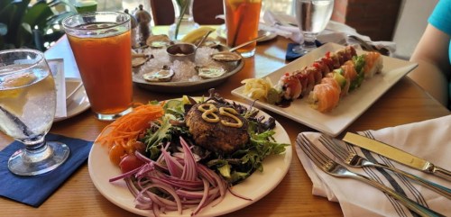 A table setting with a salad topped with a burger, sushi platter, and drinks, including a cocktail and water.