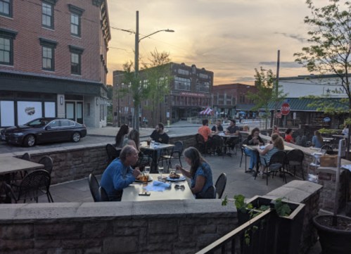 Outdoor dining scene at sunset, with people enjoying meals at tables on a street lined with buildings.
