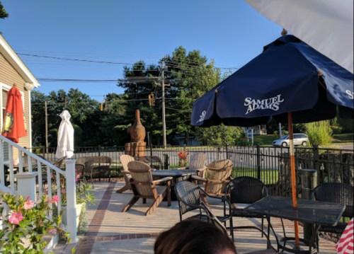 Outdoor patio with seating, umbrellas, and a wooden statue, surrounded by greenery and a clear blue sky.
