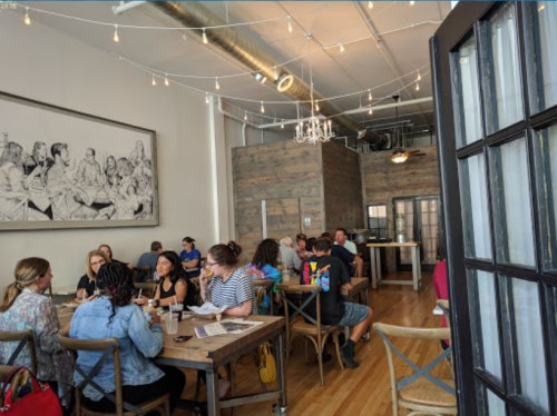 A bustling restaurant interior with diners at wooden tables, string lights overhead, and a large artwork on the wall.