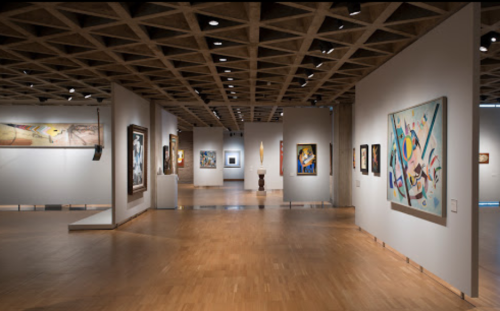 A spacious art gallery featuring various paintings and sculptures displayed on white walls and wooden floors.