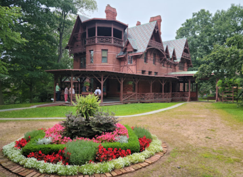 A large, historic wooden house with intricate architecture, surrounded by a colorful flower garden and green trees.