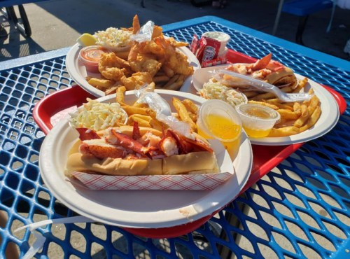 Three trays of seafood dishes featuring lobster rolls, fried shrimp, coleslaw, and fries on a blue table.