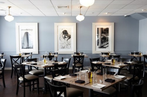 A modern restaurant interior with wooden tables, black chairs, and large framed black-and-white photographs on the walls.