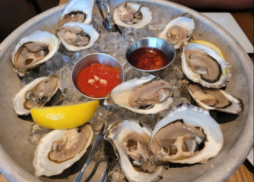 A platter of fresh oysters on ice, served with lemon wedges and two dipping sauces.