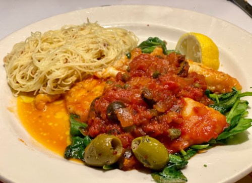 A plate of fish topped with tomato sauce and olives, served with sautéed greens and a side of pasta and lemon wedge.