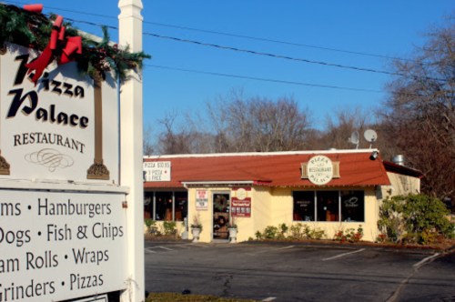 A cozy restaurant named Pizza Palace with a red roof, featuring a sign and a parking lot in a wooded area.