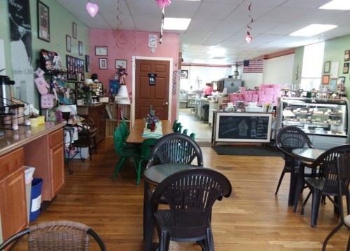 Cozy café interior with green chairs, wooden tables, and a display case filled with desserts. Pink and green walls.