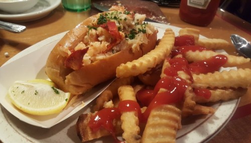 A lobster roll with lemon and a side of crinkle-cut fries drizzled with ketchup on a plate.