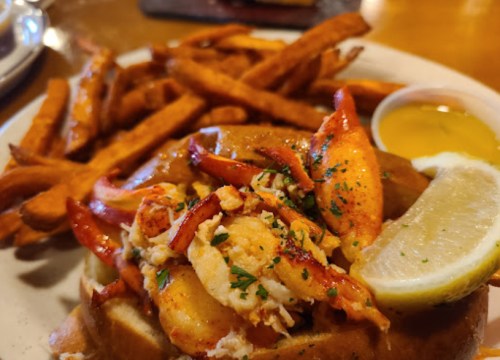 A plate featuring a sandwich topped with shrimp and crab, served with sweet potato fries and a lemon wedge.