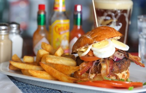 A close-up of a burger topped with bacon, egg, and vegetables, served with fries and condiments in the background.