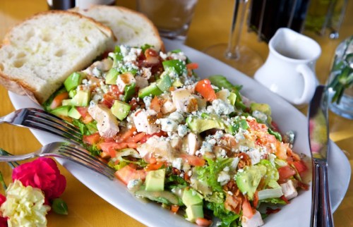A colorful salad with chicken, avocado, tomatoes, and cheese, served with two slices of bread on a yellow tablecloth.