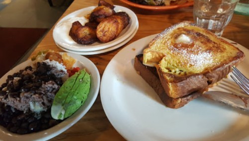 A plate with French toast dusted with powdered sugar, alongside a side of rice, beans, and fried plantains.