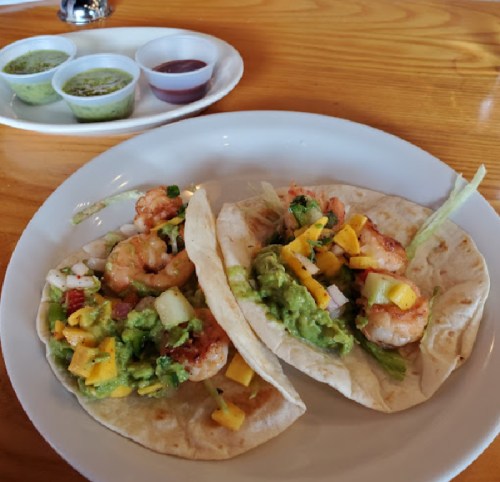 Two shrimp tacos topped with guacamole, mango, and vegetables, served with three dipping sauces in the background.