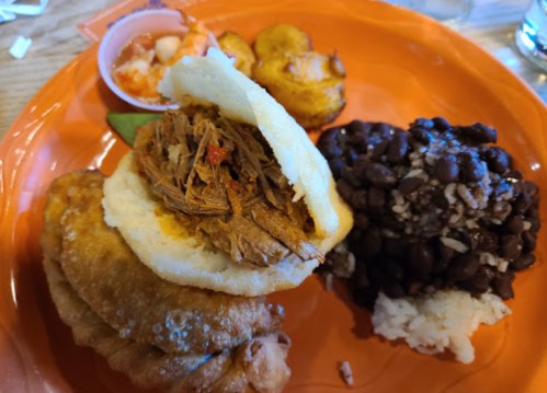 A colorful plate featuring arepas with shredded meat, fried plantains, black beans, and a small cup of shrimp.