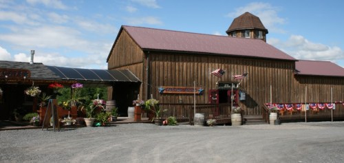 A rustic wooden barn with a red roof, surrounded by flower pots and American flags, under a blue sky.