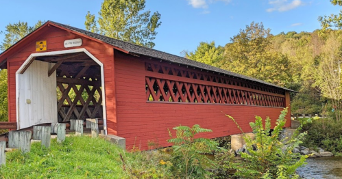 A red covered bridge spans a stream, surrounded by green trees and a clear blue sky.