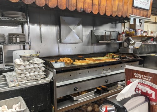 A busy kitchen with a large griddle, cooking various dishes, and shelves stocked with plates and eggs.