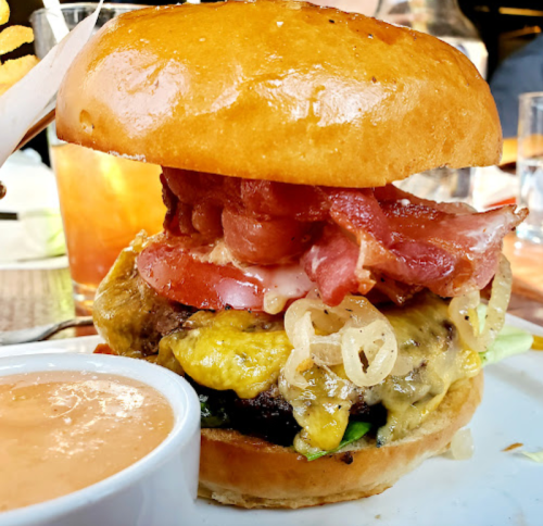 A close-up of a juicy burger with bacon, cheese, onions, and a glossy bun, served with a side of dipping sauce.