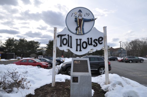 Sign for Toll House with a figure pointing, surrounded by snow and parked cars, under a cloudy sky. Established 1709.