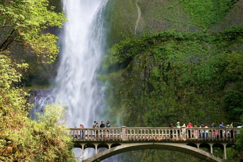 A stone bridge with people standing on it, overlooking a tall waterfall surrounded by lush greenery.