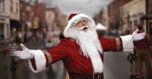A cheerful Santa Claus with a big smile and open arms stands in a festive street scene.