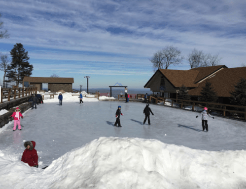 Children and adults ice skating on a rink surrounded by snow, with a lodge in the background and a clear blue sky.