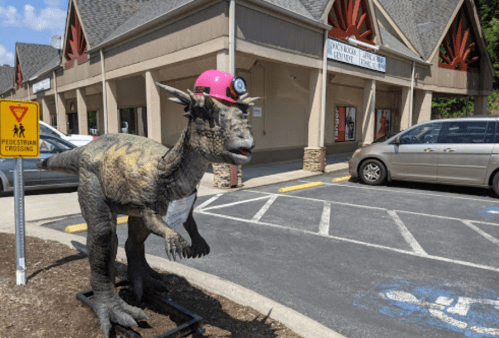 A dinosaur statue wearing a pink hard hat stands outside a building, with a parking lot and pedestrian sign nearby.